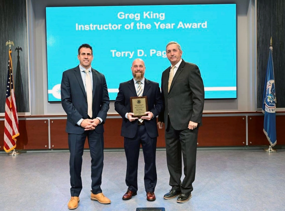 Terry Page (Center) of the Federal Law Enforcement Training Centers (FLETC), Physical Techniques Division, Core Training Operations Directorate, is presented the Greg King Instructor of the Year award for 2023, by FLETC Benjamine C. Huffman (Right) and FLETC Deputy Director Kai J. Munshi (Right) at the FLETC Annual Awards Ceremony, June 12, 2024, Glynco Ga. (Photo by David Tucker, OPA)